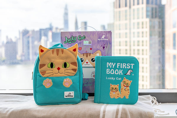 A product shot of the My First Book Lucky Cat busy book set against a city skyline, featuring the Montessori-inspired busy book, a waterproof cat-themed backpack, and gift-ready packaging—making it a perfect baby gift.