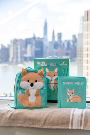 A product shot of the My First Book Nordic Forest busy book set against a city skyline, featuring the Montessori-inspired busy book, a waterproof fox backpack, and gift-ready packaging—an ideal baby gift.