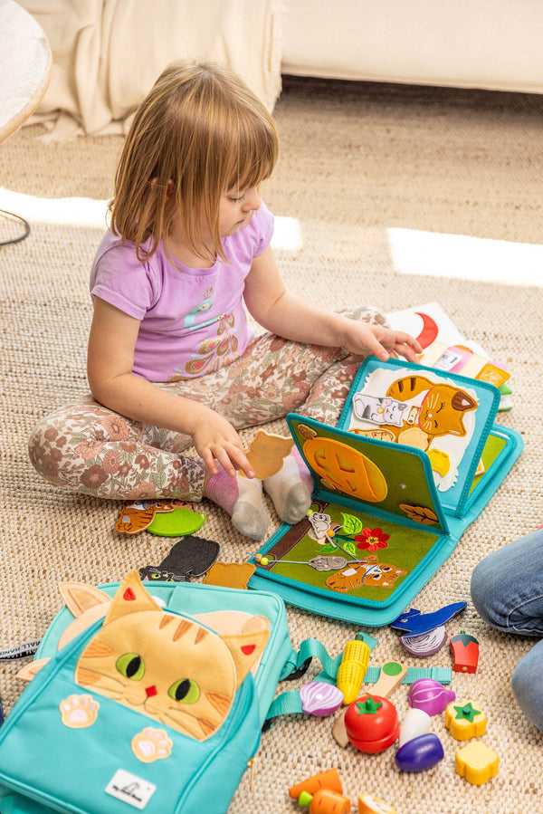 A serene scene of a girl quietly exploring the My First Book Lucky Cat busy book, with sensory features and Montessori-inspired design, her cat-themed backpack resting by her side. An ideal gift for babies.