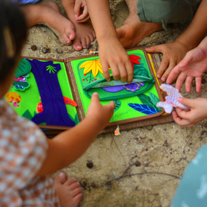 A lively scene with multiple kids gathered on the beach, engrossed in the My First Book Dinosaur busy book. They explore the interactive, dinosaur-themed pages together, building sensory skills through Montessori-inspired play in a fun, outdoor setting.