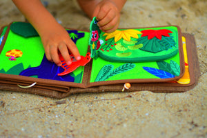 Picture shows a kid playing dinosaur busy book on beach, showing the high-quality details and sensory interactive play in this Montessori inspired book. Makes a great baby gift.