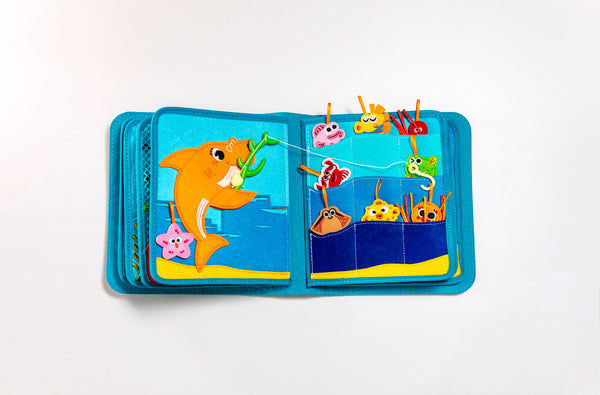 Two pages of My First Book Baby Shark busy book product photo, showing the cute characters and sensory interactive fishing game play in this Montessori inspired book. Makes a great baby gift.