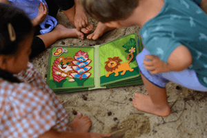 toddlers playing with MyFirstBook fabric busy book featuring colorful tiger and lion designs on sand